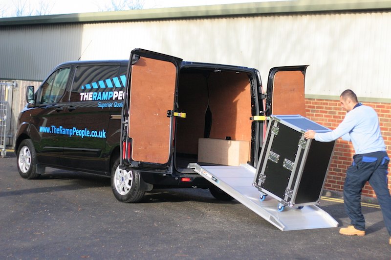 Van with open doors and a ramp, being loaded with a large black flight case by a person.