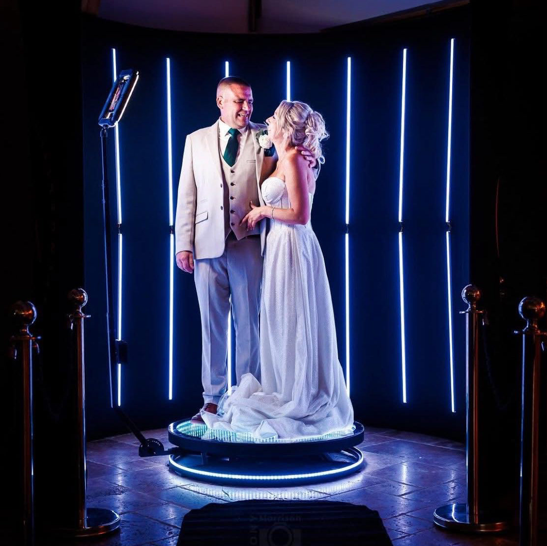 Man and woman standing on a circular photobooth with blue lights against a dark background