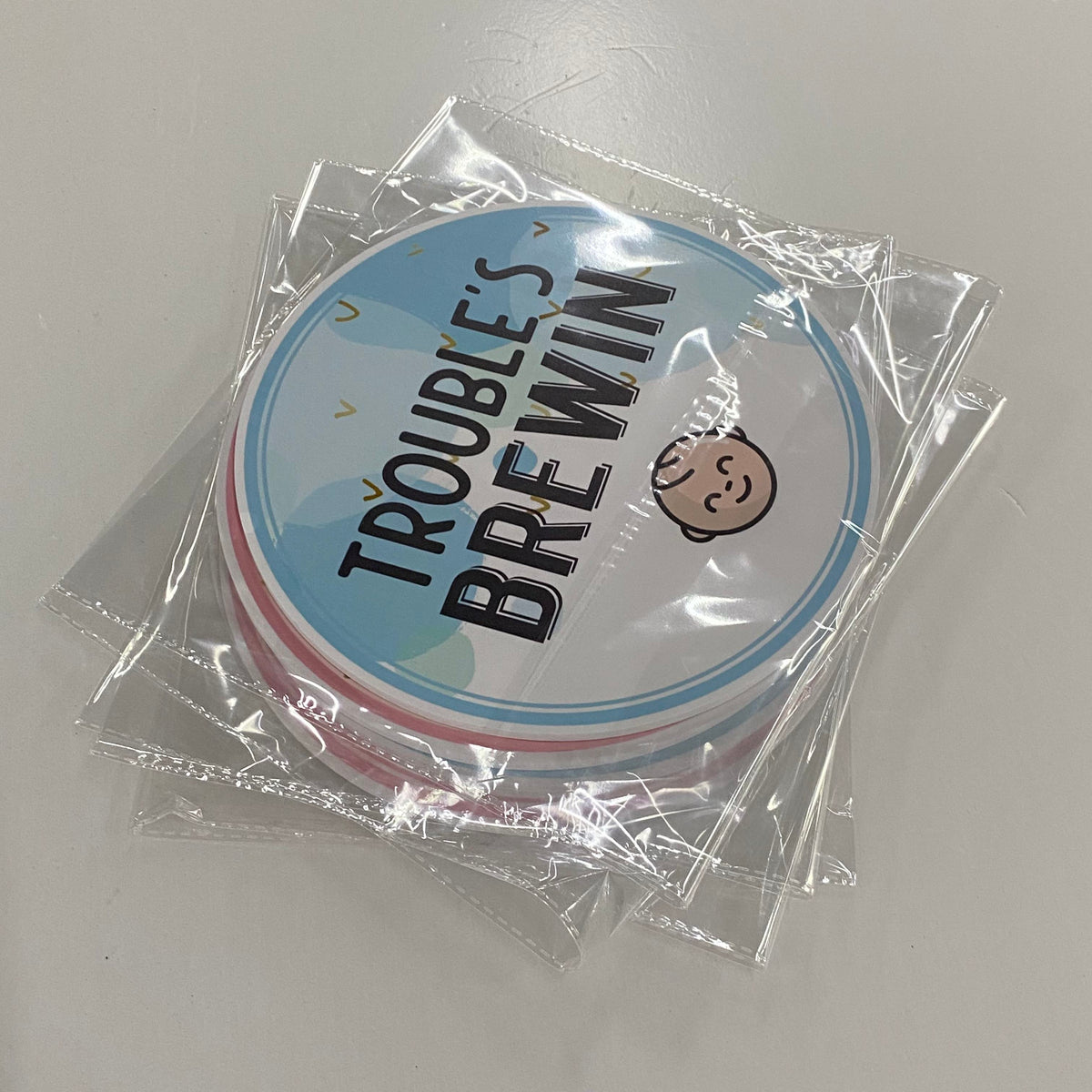 Round photobooth prop with 'Trouble's Brewin' text and baby graphic in clear packaging on a gray background