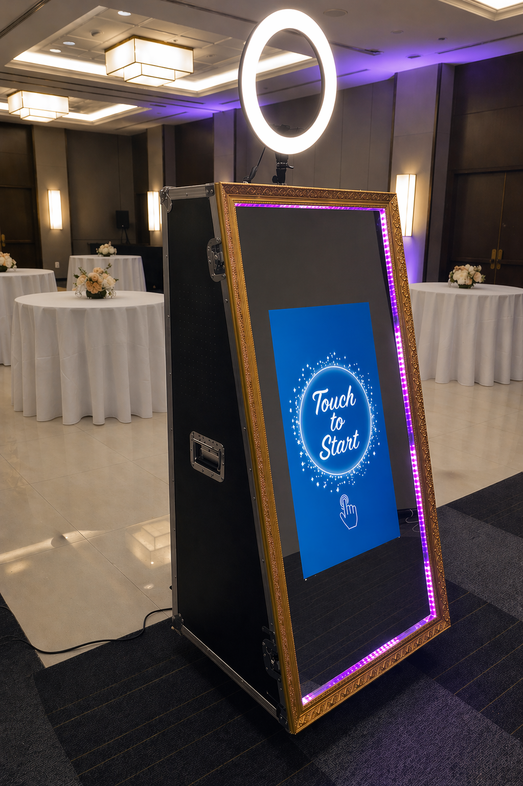 Interactive touch screen magic mirror photobooth with a blue screen displaying 'Touch to Start' in a room with tables and chairs.