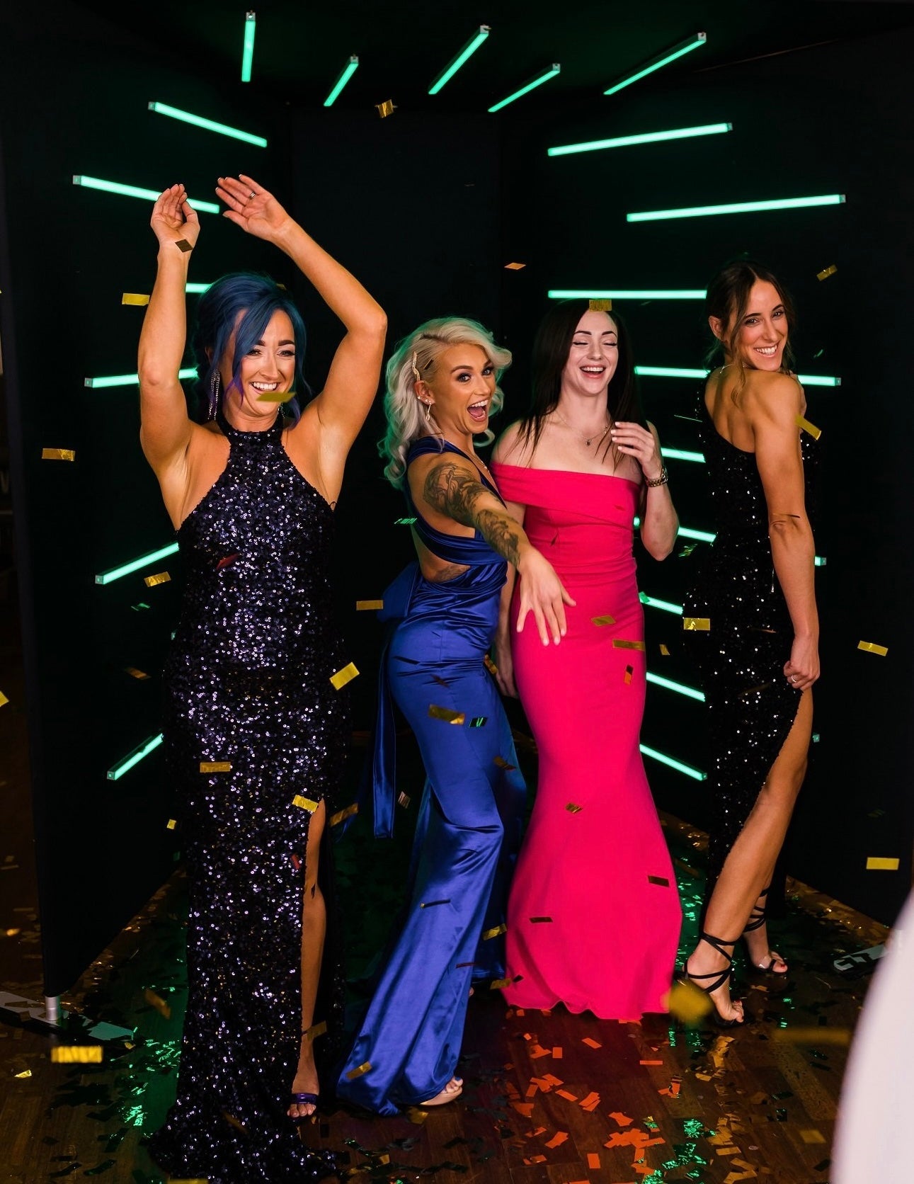 Four women in glamorous outfits posing together with a neon light background