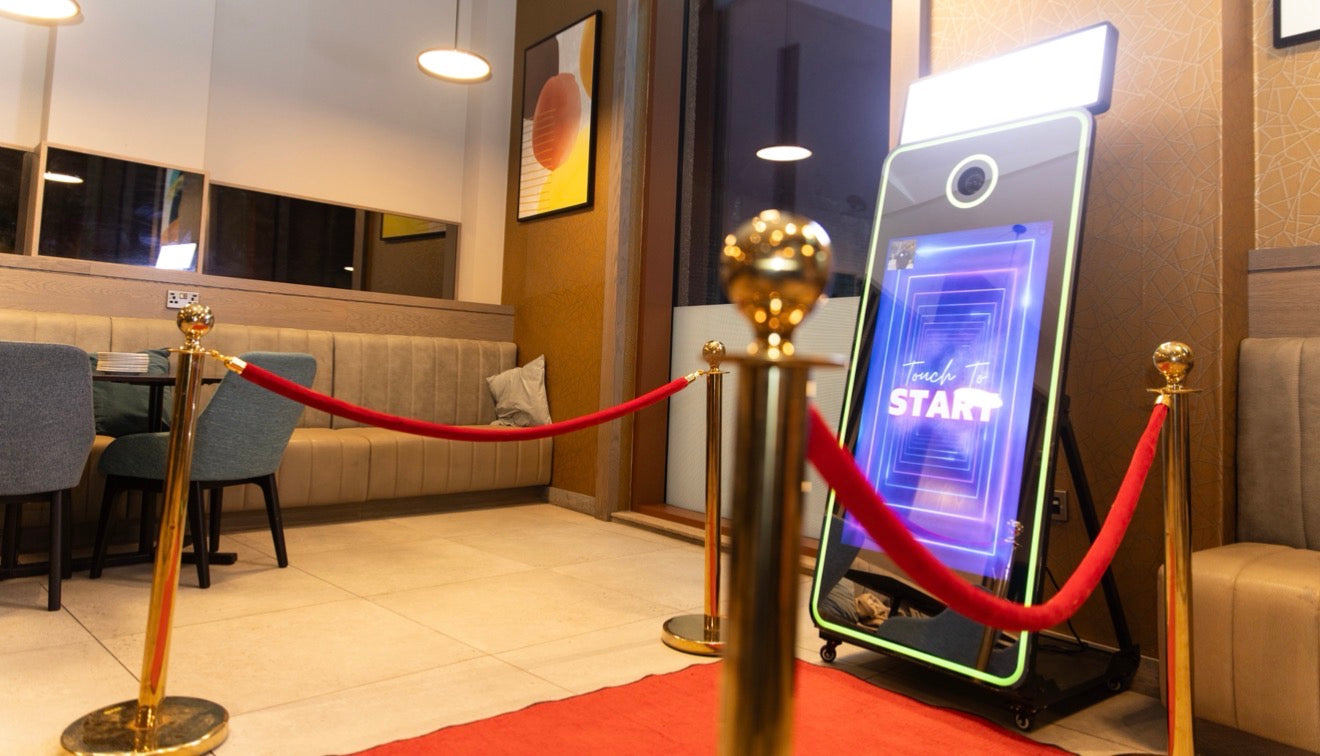 Interactive touch screen photobooth in a modern restaurant setting with red velvet rope and gold posts.