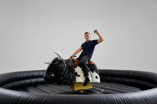 Man riding a black mechanical bull with a round black inflatable support. 