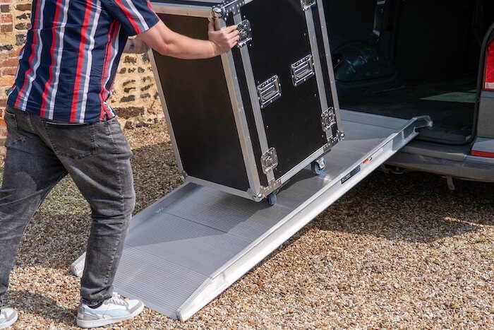 Person using a large, sturdy aluminium ramp to load a large black flight case into a vehicle