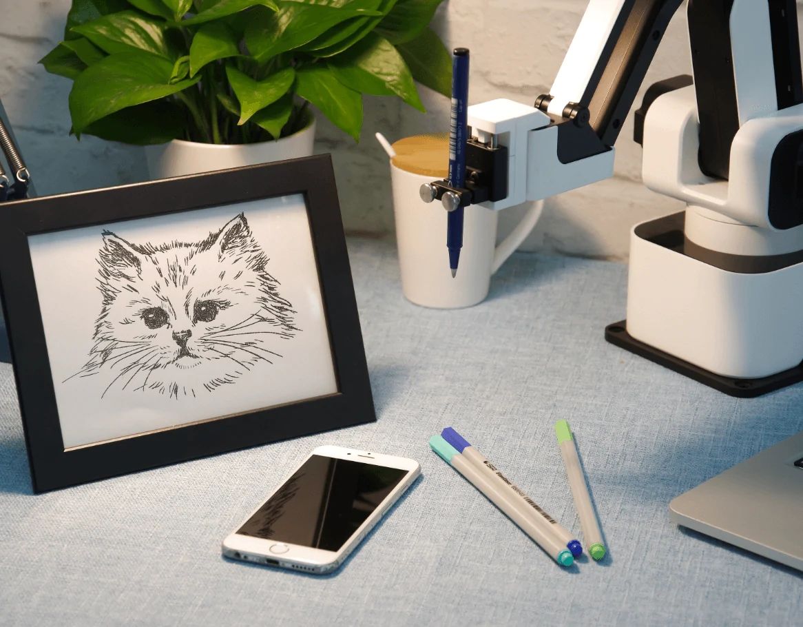Desk setup with a framed drawing of a cat, with the robot arm that created the drawing.
